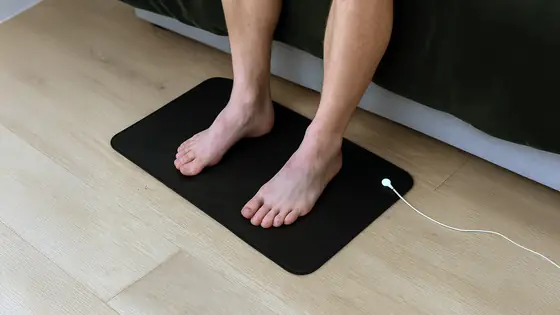 Official image of bare feet resting on the Velin Grounding Mat beside a bed
